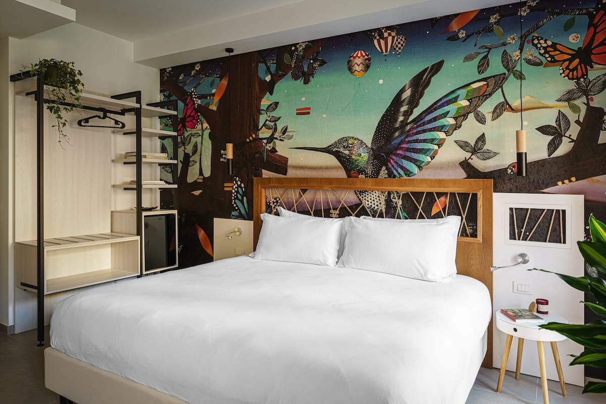 Un dormitorio con cama, mesita de noche, una pequeña mesa auxiliar, una planta colgante y un mural en la pared.
