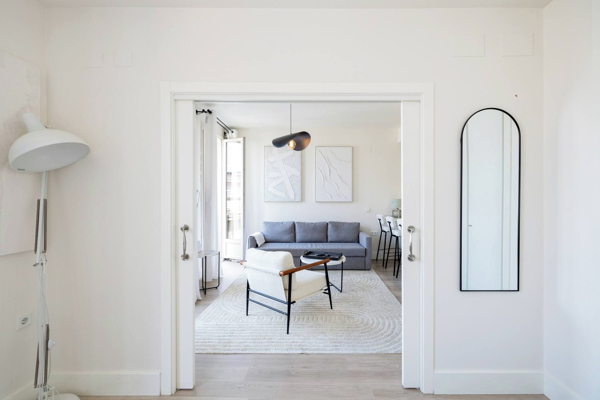 Una sala de estar moderna con un sofá gris, un sillón blanco y una mesa de centro. Hay un espejo grande en la pared y una lámpara de pie.