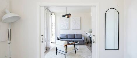 Un salon moderne avec un canapé gris, un fauteuil blanc et une table basse. Un grand miroir orne le mur, et une lampe sur pied se trouve dans la pièce.