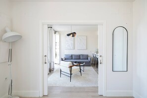 Een moderne woonkamer met een grijze bank, een witte fauteuil en een salontafel. Er hangt een grote spiegel aan de muur en staat een vloerlamp.