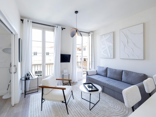 Ein modernes Wohnzimmer mit grauem Sofa, einem weißen Sessel und einem Couchtisch. An der Wand ist ein Fernseher angebracht und es gibt ein Fenster mit Vorhängen.