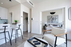 Un salon moderne avec un canapé, une table basse et un téléviseur. Il y a un coin cuisine équipé d’un réfrigérateur, d’une cuisinière et d’un évier. La pièce est dotée d’une grande fenêtre et d’une porte qui donne sur une autre pièce.