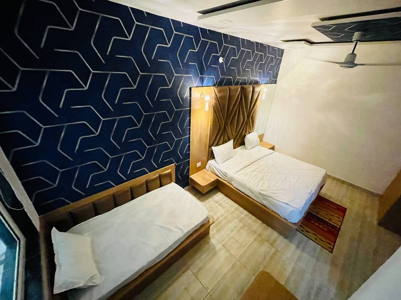Habitación de hotel con dos camas individuales, un papel tapiz estampado, un ventilador de techo y un cabecero de madera.