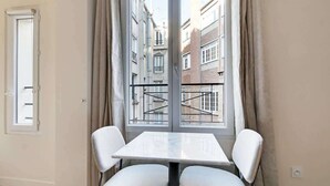Ein Zimmer mit Tisch und zwei Stühlen, einem fenster mit Vorhängen und Blick auf Gebäude.