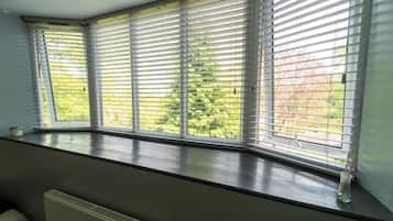 Quarto com janela saliente com persianas horizontais, radiador e vista para o verde lá fora.