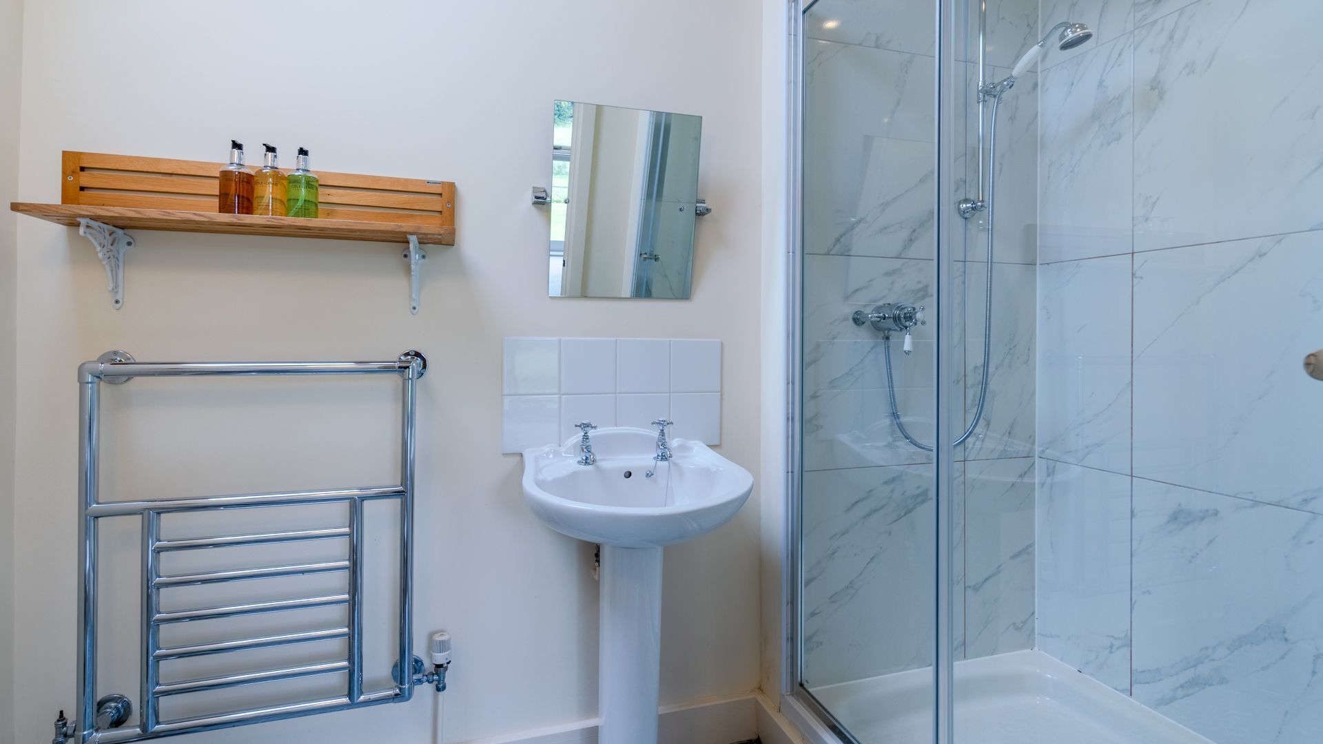 Une salle de bain avec une douche à parois de verre, un lavabo sur pied blanc et une étagère en bois où sont rangés les articles de toilette.