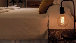 Un lit bien fait, avec du linge de lit blanc et un seul oreiller jaune. Une lampe de chevet moderne, dotée d’un abat-jour en verre transparent, est posée sur la table de chevet.
