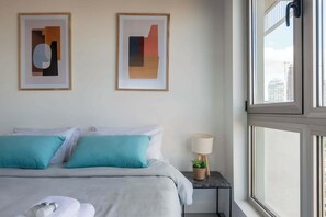 Habitación de hotel con cama, dos almohadas, mesita de noche con lámpara y dos cuadros abstractos en la pared.