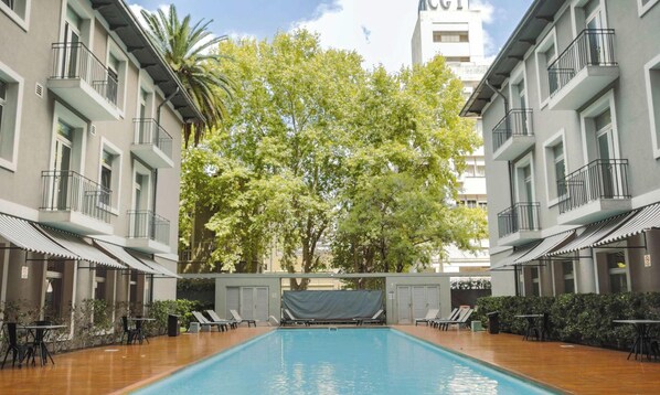 Una piscina con sillas y mesas de descanso, rodeada de edificios con balcones y árboles.