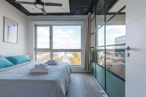 Una habitación de hotel moderna con una cama grande, un ventilador de techo y vistas a la ciudad a través de amplios ventanales.