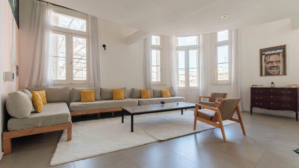 A modern living room with a sectional sofa, a coffee table, and a wooden chair. There are large windows with curtains, a dark wooden cabinet, and a poster on the wall.