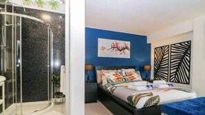 A modern bedroom with a shower, a bed with floral bedding, a blue accent wall, and a decorative leaf pattern on the wall. - English Riviera Coastal Holiday Home in Devon Uk (Paignton)