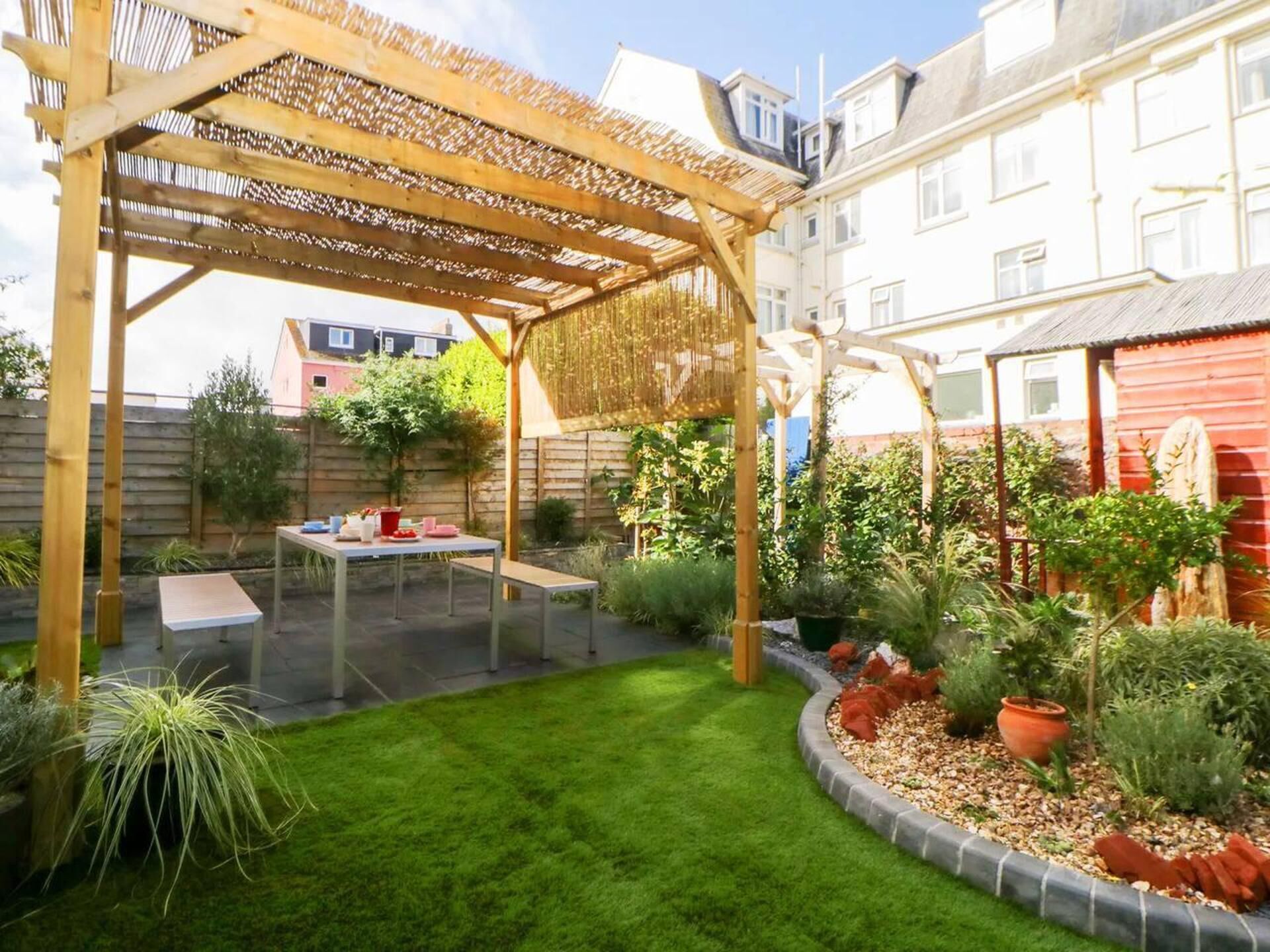 A covered patio area with a table and benches, surrounded by a well-maintained lawn and various plants.