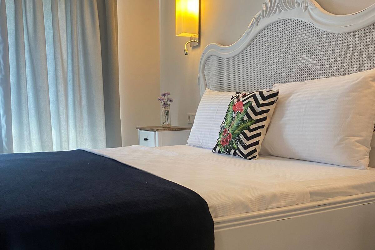 A neatly made bed with a black throw blanket, a white headboard, and a wall-mounted light fixture.