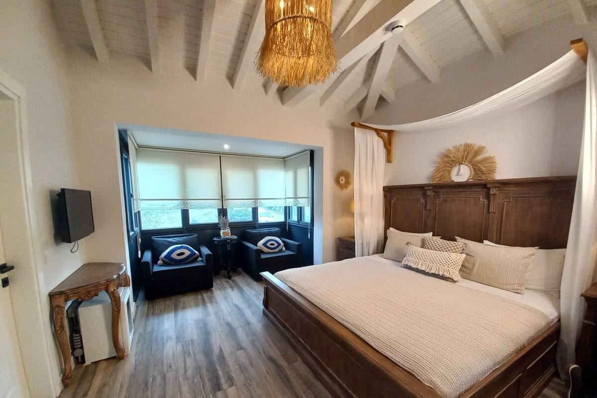 A bedroom with a large bed, wooden headboard, and a canopy. There are two armchairs and a small table with decorative pillows.