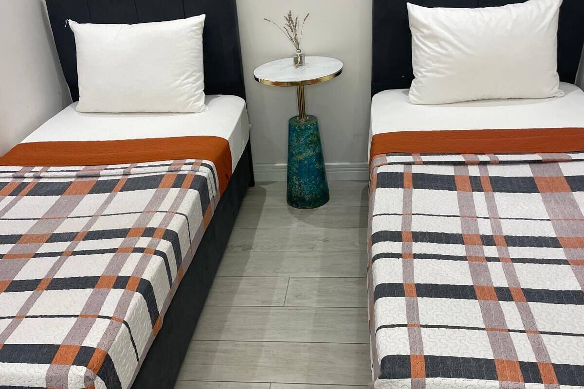 A hotel room with two beds, each with a white pillow and a checkered blanket. There is a small round table with a vase of dried flowers between the beds.