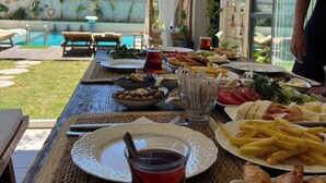 A dining table with various dishes, a glass of red drink, and a view of a swimming pool and buildings in the background. - Chloe Alacati (Cesme)