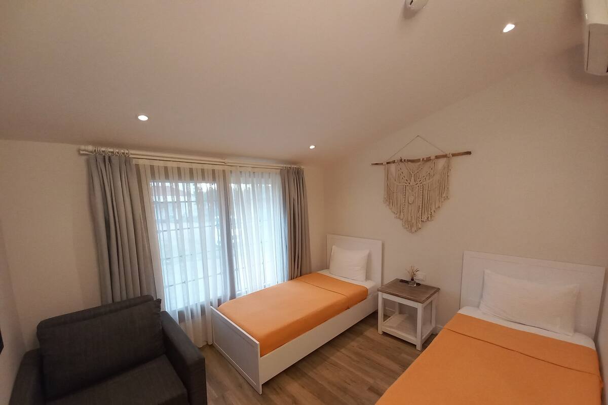 A hotel room with two beds, a small table, a chair, and a macrame wall hanging.