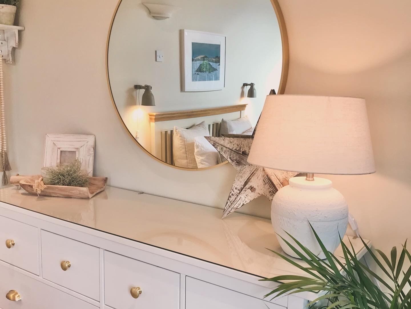 Une pièce bien éclairée, avec un grand miroir rond, une lampe de chevet, une table de chevet ornée d’une plante et un lit doté d’une tête de lit.