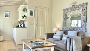 A living room with a sofa, a coffee table, a mirror, and a kitchen area in the background. - Adults-Only Waterfront Guest Suite | Stunning Harbour Views | Cozy Fireplace (Kinsale)