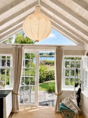 A room with a large window, a hanging light fixture, and a view of a garden and water body.