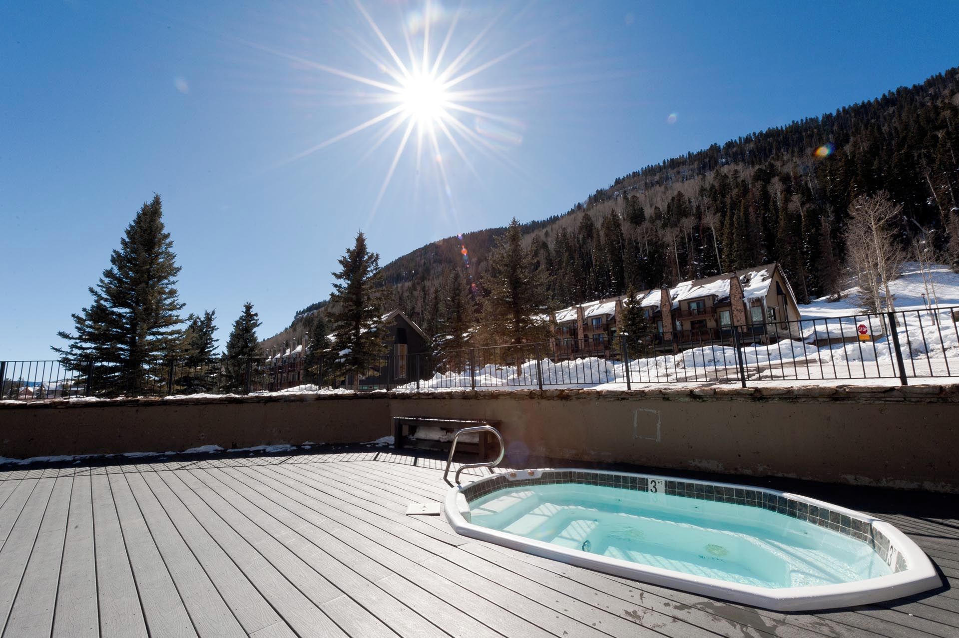 A wooden deck with a hot tub, surrounded by snow-covered ground and a mountainous backdrop.