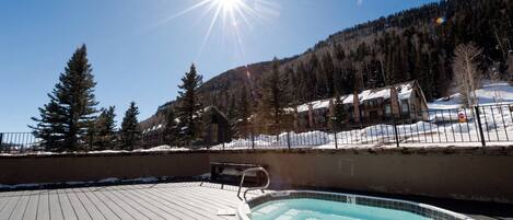 A wooden deck with a hot tub, surrounded by snow-covered ground and a mountainous backdrop.