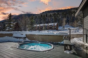 A wooden deck with a hot tub, a bench, and a view of a lodge in a snowy mountainous area.