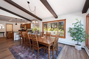 Un comedor amplio con una mesa y sillas de madera, un ventanal con vistas a árboles, y una cocina al fondo.