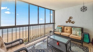 Un balcón con vista a la playa, un sofá de mimbre con cojines, una mesita y una lámpara colgante.