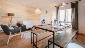 A modern living room with a wooden floor, a dining table set for four, a sofa, a lamp, and a wall-mounted deer head decoration.