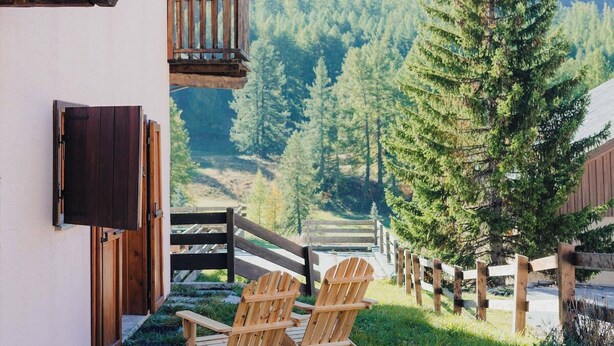 A chalet with a wooden balcony, two Adirondack chairs, and a mountain view.