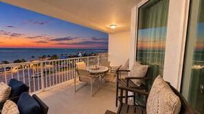 A balcony with a view of the ocean at sunset, furnished with a table and chairs, and a sofa with cushions.