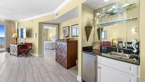 Une chambre d’hôtel avec un grand lit, un coin salon avec un fauteuil rouge, une cuisine équipée d’un évier et de placards, et un grand miroir agrémenté d’objets décoratifs.