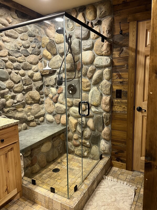 A stone-walled shower enclosure with a glass door, wooden cabinetry, and a rustic wooden door.