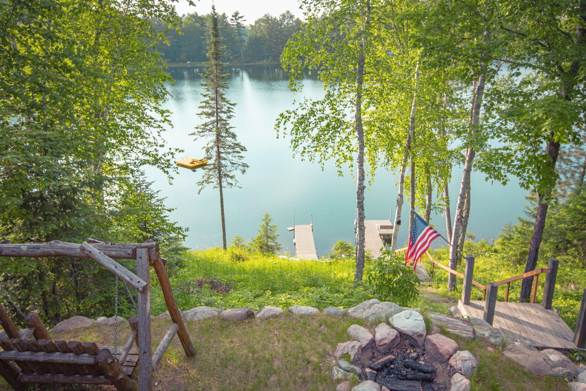 A lakeside cabin with a deck, a fire pit, and an American flag.