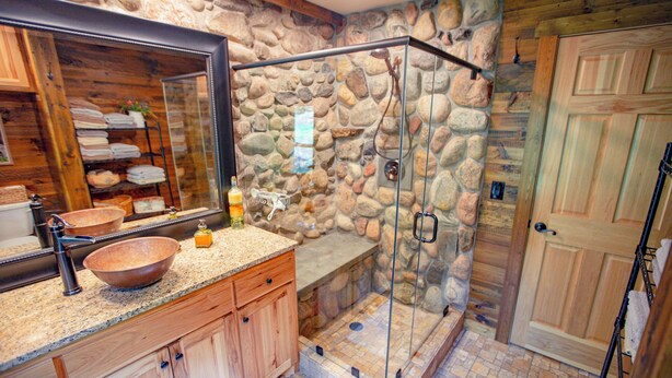 A bathroom with a stone wall, a large mirror, a wooden vanity, a glass shower enclosure, and a wooden door. - Cozy Stone Cottage overlooking Smith Lake (Marcell)