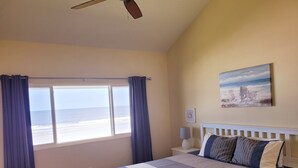 A bedroom with a bed, a ceiling fan, a window with curtains, a nightstand with a lamp, and a painting on the wall.
