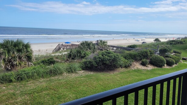 A beach view with a wooden deck, grassy area, and a clear sky.
