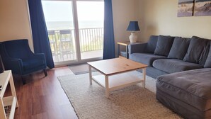 A living room with a sofa, a table, and a chair. There is a view of the outdoors through a glass door.