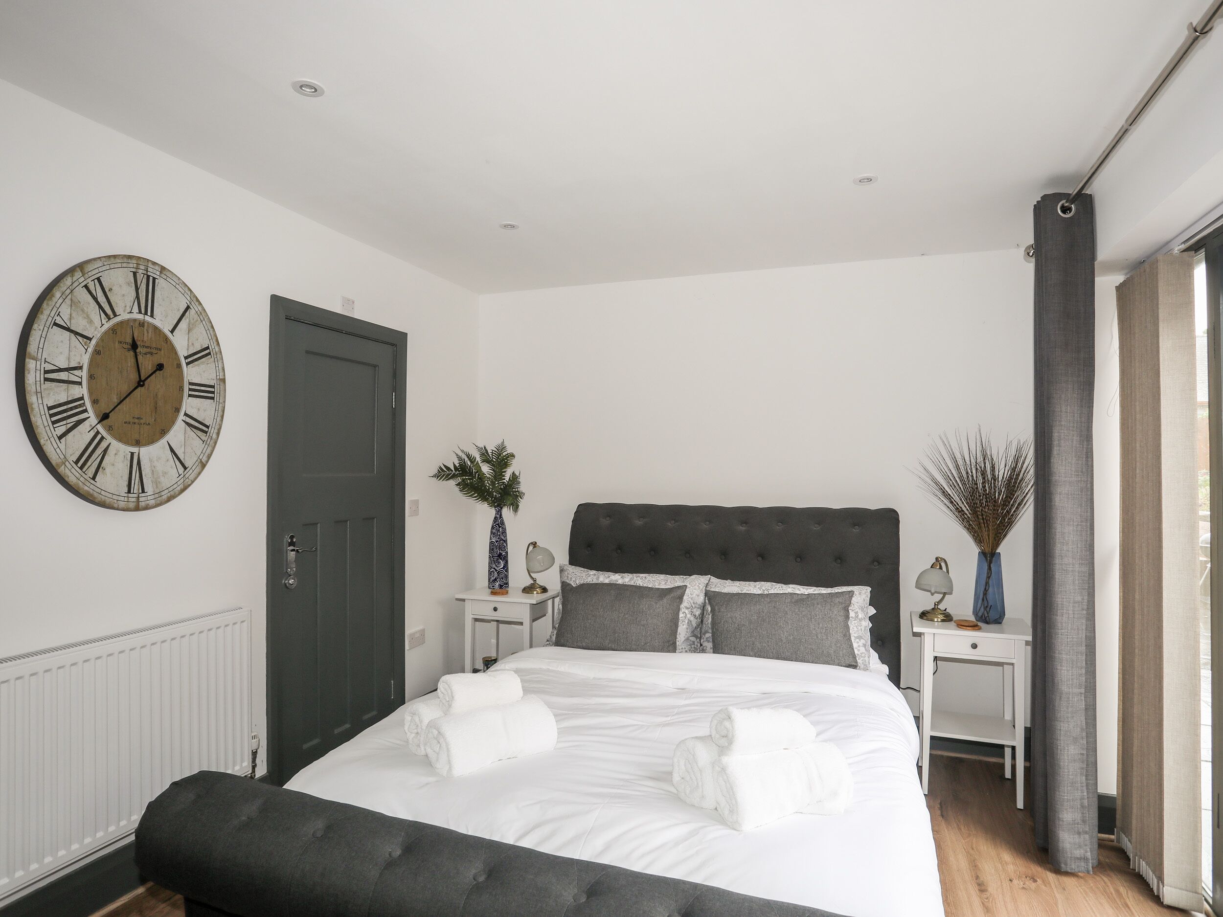 Een slaapkamer met een grote wandklok, een bed met witte lakens, een donkere hoofdeinde en een raam met gordijnen.