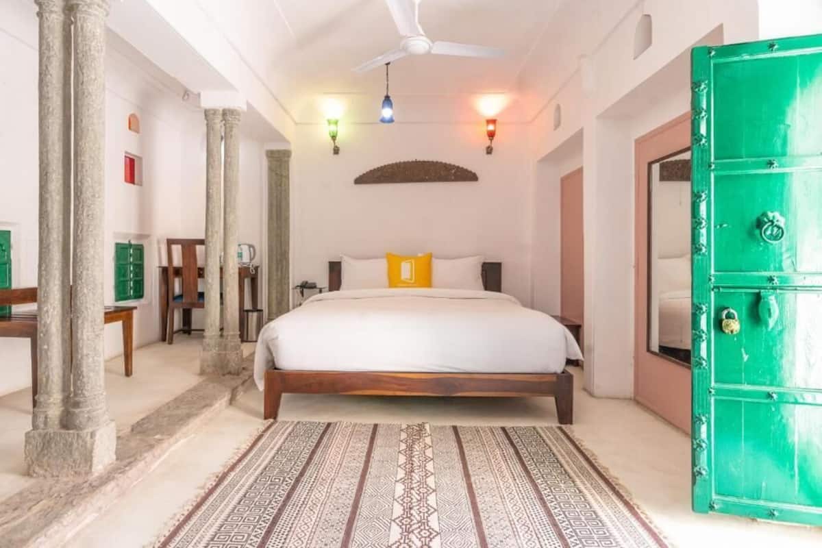 Um quarto com uma cama, uma porta verde, um ventilador de teto e um tapete estampado.