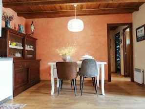 Zona pranzo con tavolo bianco, due sedie, un vaso con piante essiccate, un mobiletto in legno e un lampadario.