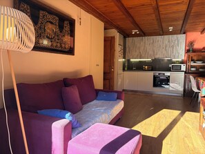 A living room with a sofa, a small table, a lamp, a kitchen area with appliances, and a wooden ceiling. - Ground floor with garden and barbecue - Puig Falcó 2 (Pla de l'Ermita)