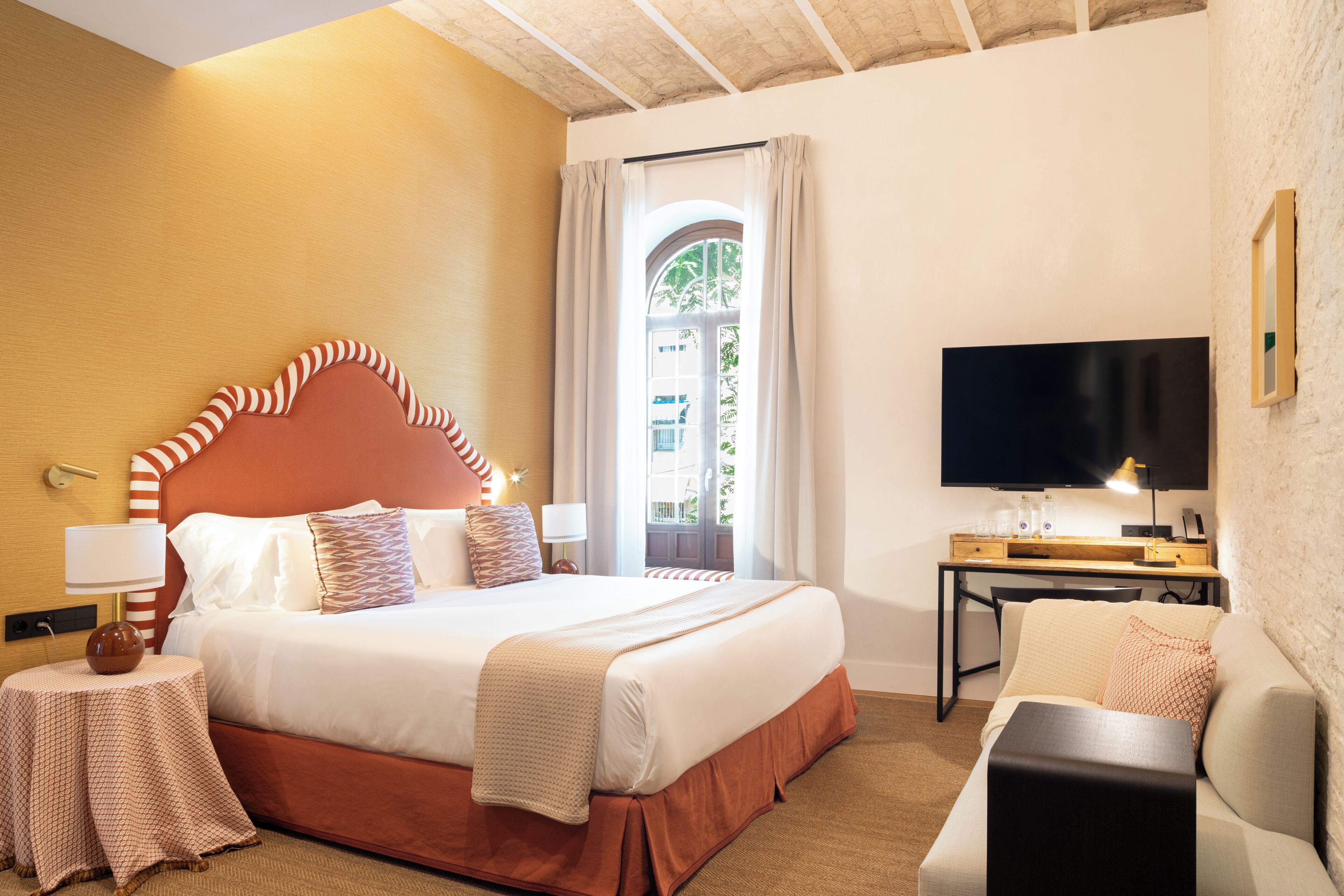 Habitación de hotel con una cama grande, un escritorio con televisor, un sofá y vista a un edificio desde la ventana.