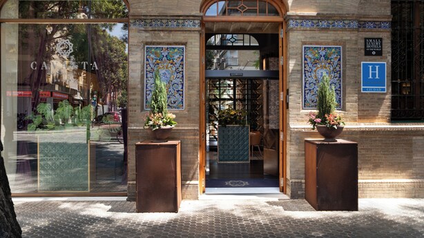 Entrada de hotel con fachada de azulejos, un letrero azul con una 'H' que indica el hotel, y dos macetas con plantas a la entrada.