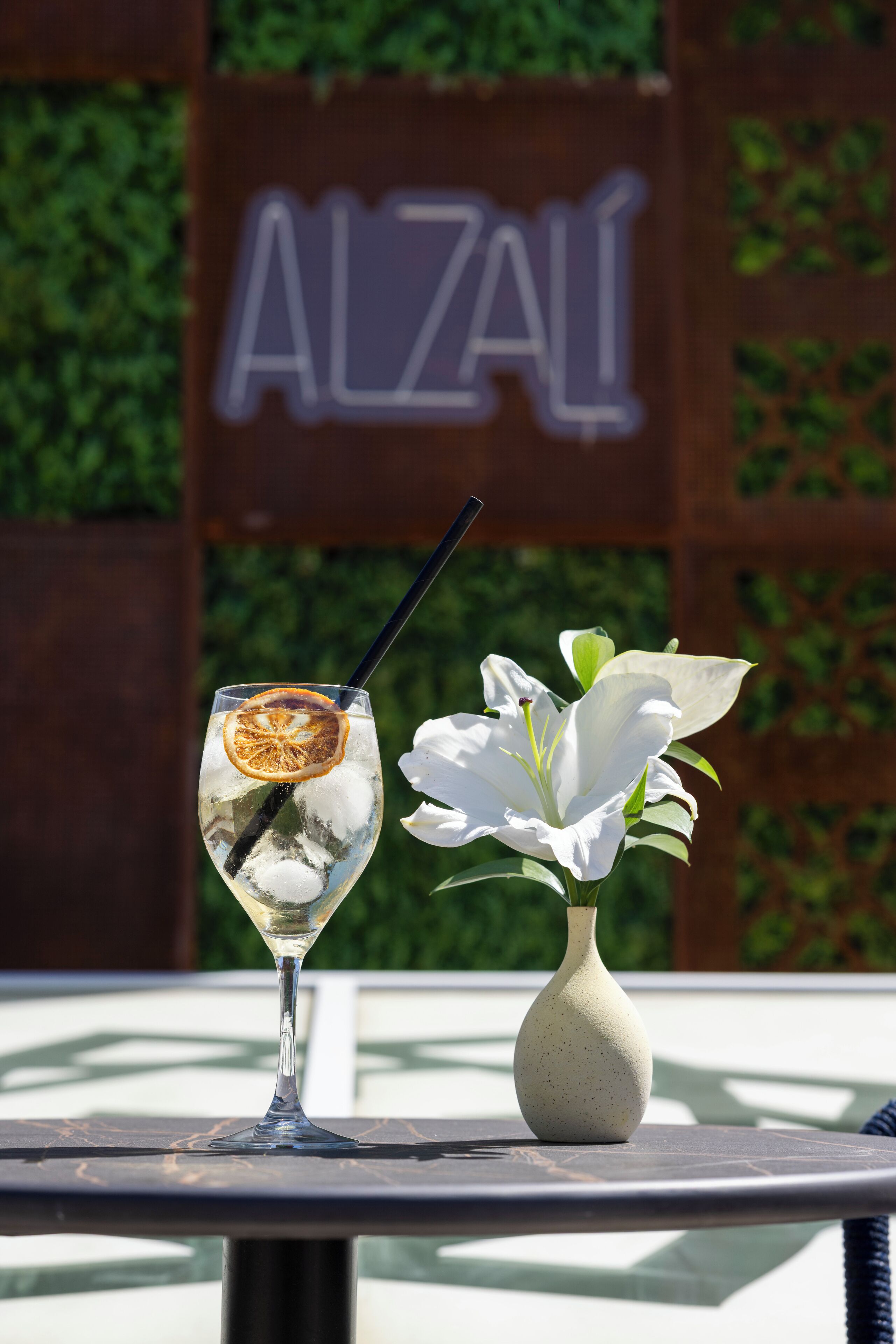 A table with a glass of a clear beverage, a dried orange slice, and a white flower in a vase.