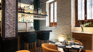 Zona de bar con pared de piedra, mostrador de bar de madera y varias botellas y vasos. Hay dos taburetes de bar y una mesa con platos y cubiertos.