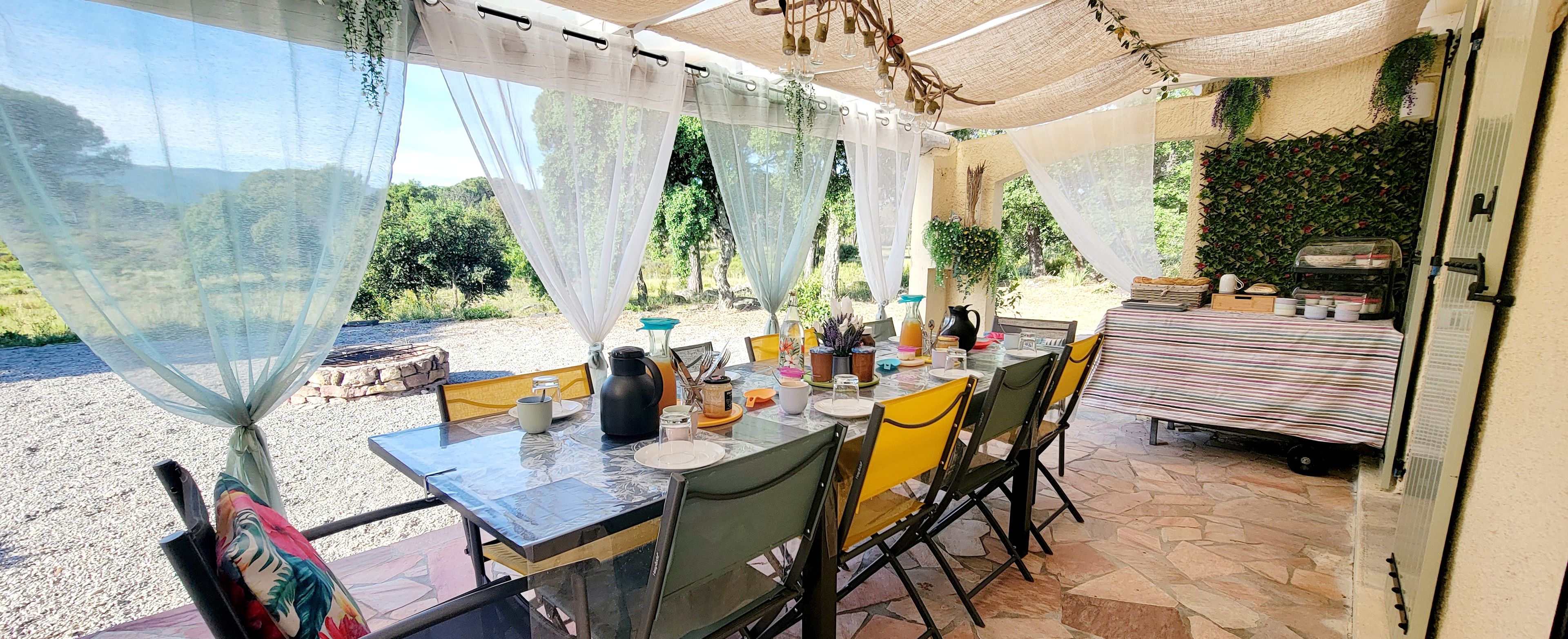Un espace extérieur aménagé pour les repas, avec une table dressée pour quatre personnes, entourée de chaises, et un barbecue à l’arrière-plan.