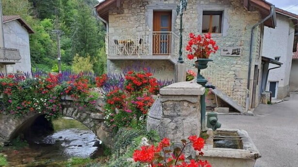 Un bâtiment en pierre doté d’un balcon, d’un petit pont enjambant un ruisseau et de jardinières ornées de fleurs rouges.